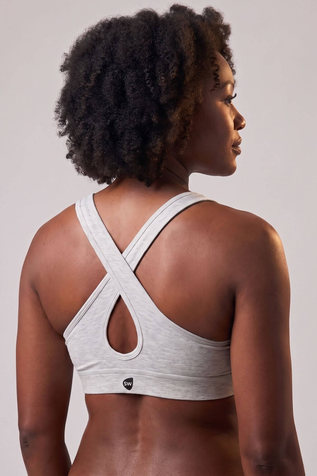 A woman shown from the back wearing a light heather gray sports bra with a crossover crisscross strap design and a keyhole cutout at the center back. The brand logo on the bottom is too small to read clearly, so no brand is listed.