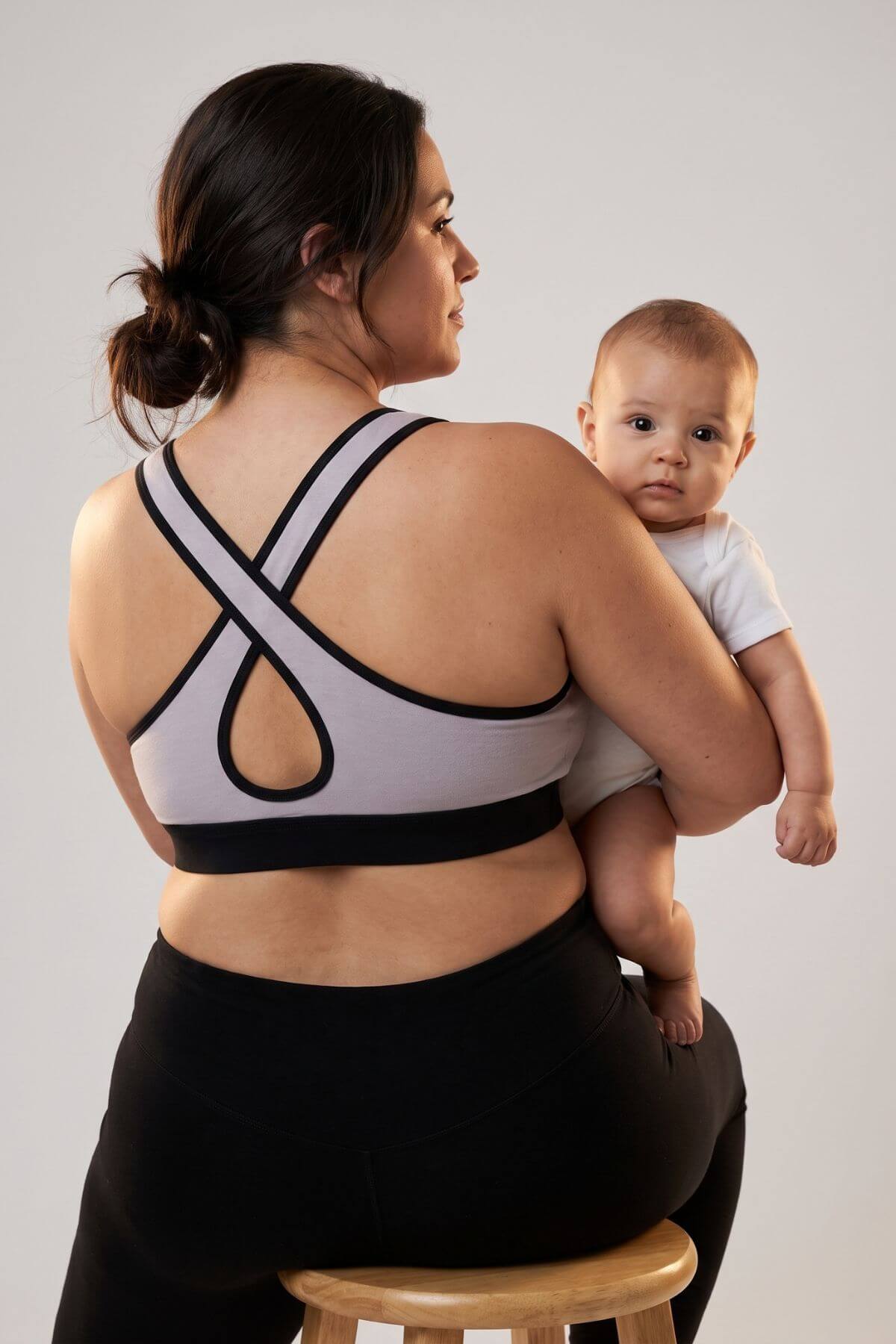 A woman wearing a light gray and black sports bra sits on a wooden stool, holding a baby dressed in a white outfit against a plain light gray background.
