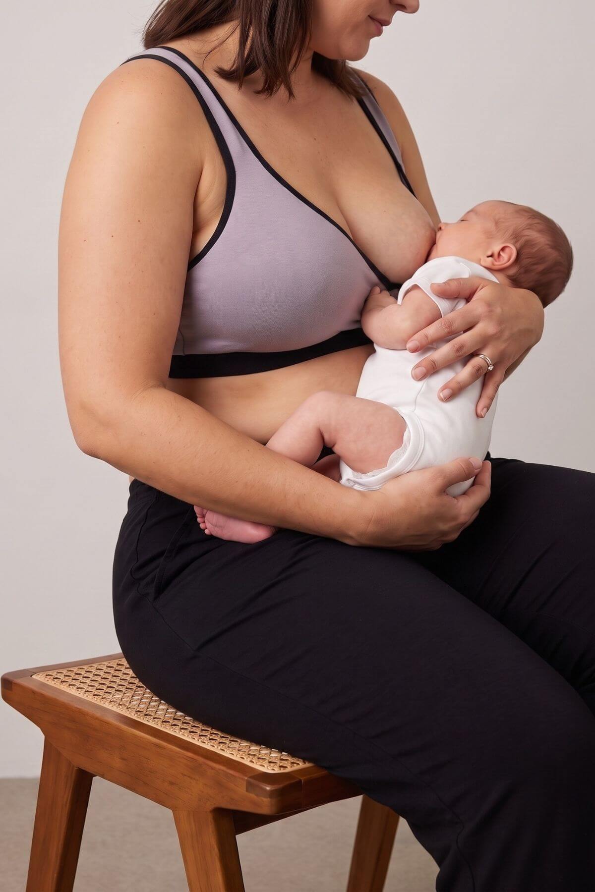 A woman in a light purple and black nursing bra holds a baby dressed in a white onesie while breastfeeding, seated on a wooden stool against a plain light gray wall.