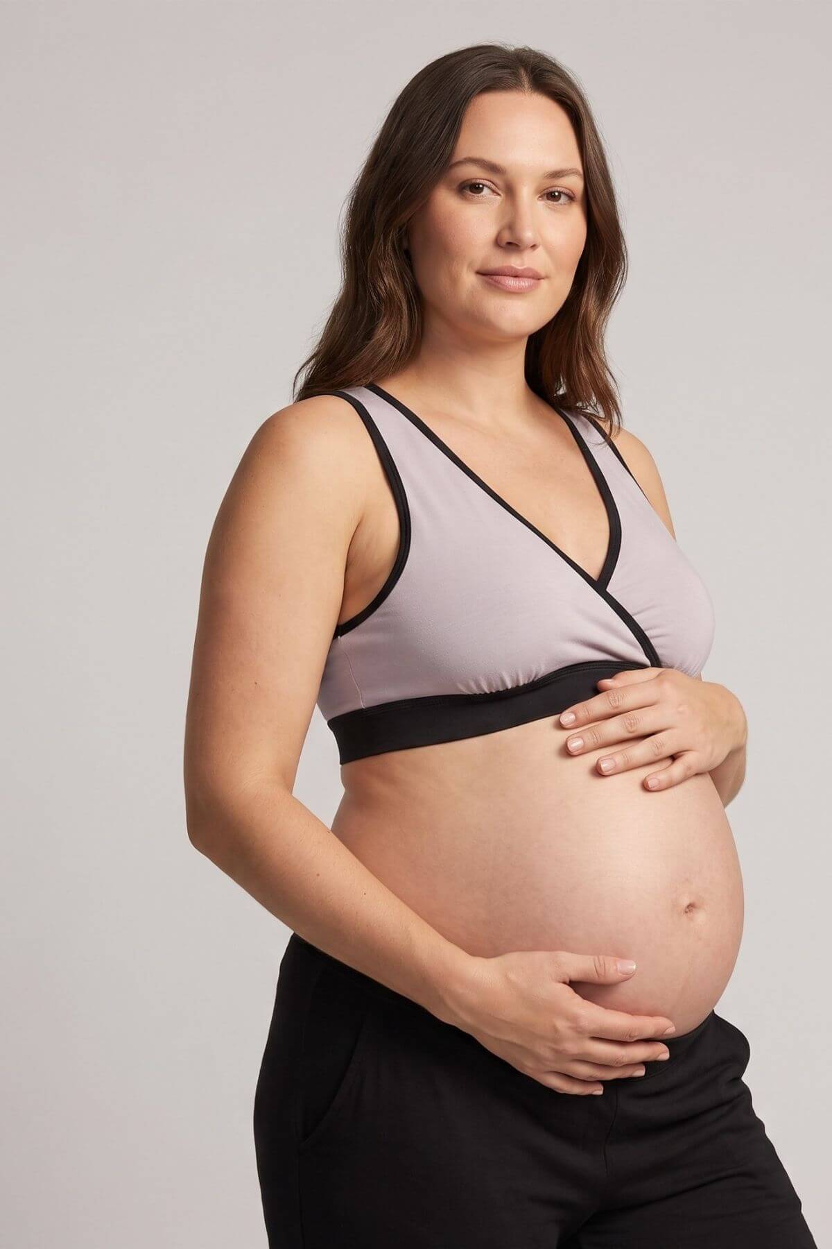 A pregnant woman wearing a light gray wrap-style maternity bra with black trim and black pants, standing against a neutral light gray background.