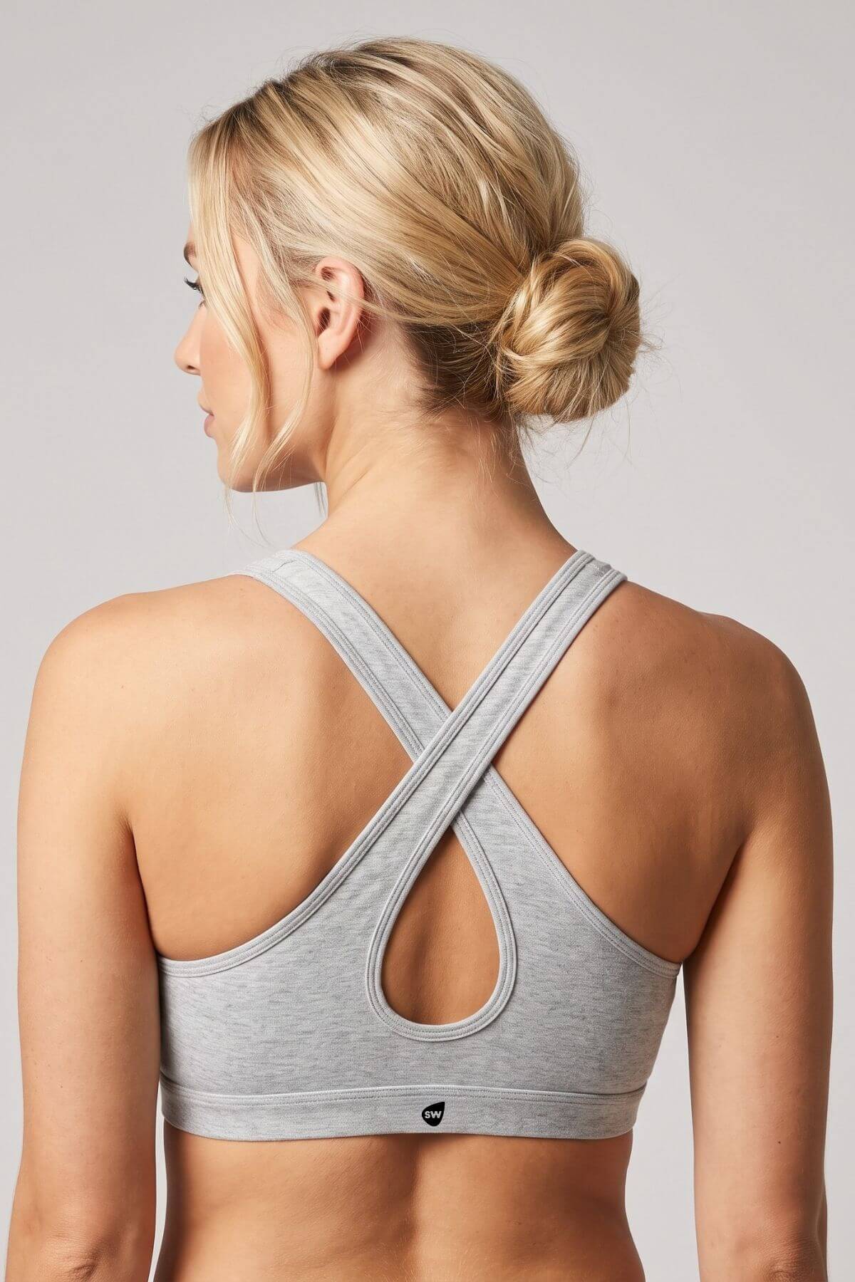 A woman shown from the back wearing a light gray heather sports bra with a crossover crisscross back strap design and a small black logo at the bottom center.