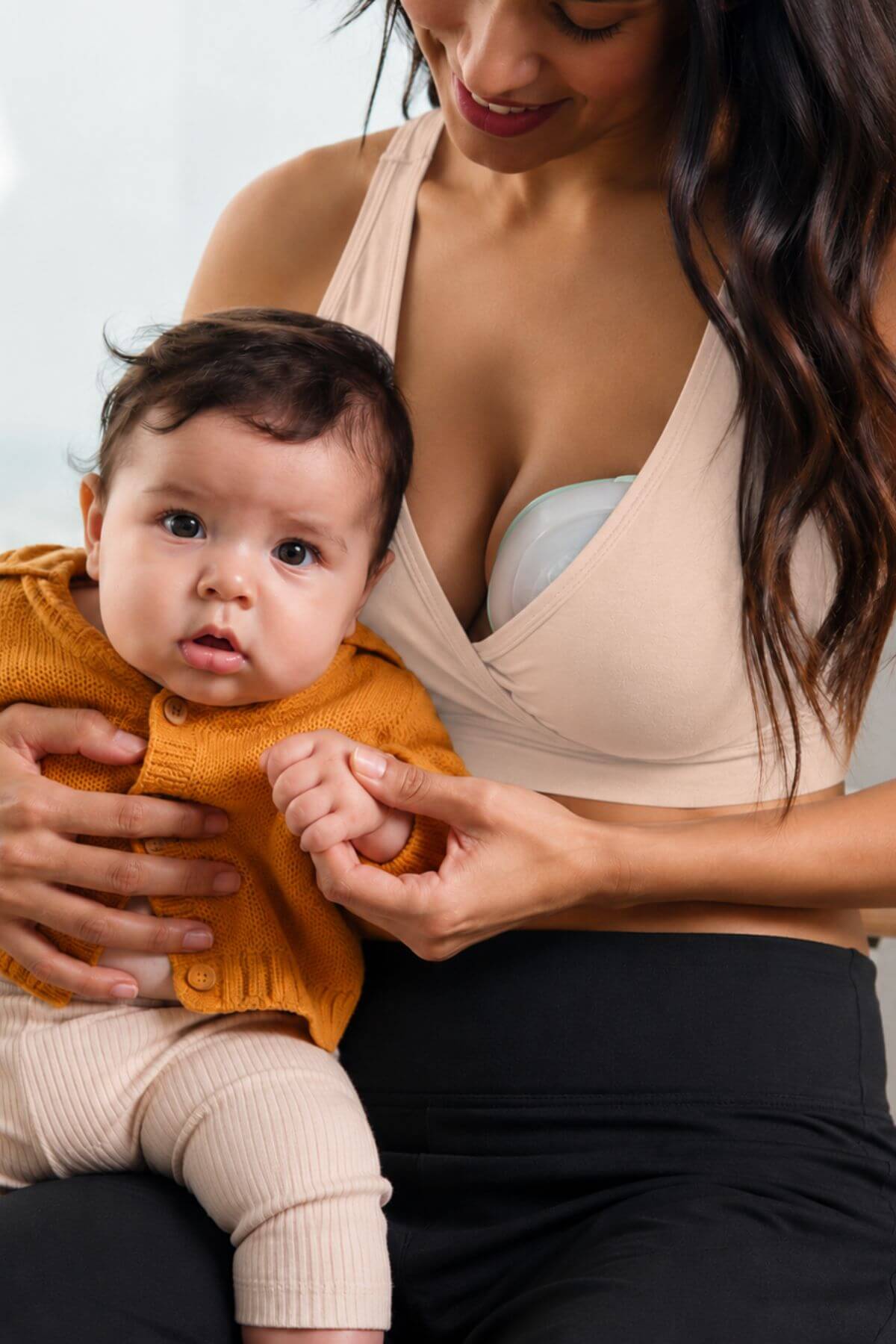 A woman wearing a beige nursing bra holds a baby dressed in a mustard yellow knit top and cream pants. The woman is adjusting the baby&