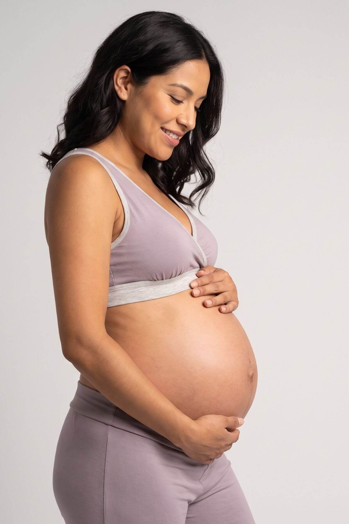 A pregnant woman wearing a light purple maternity bra and matching leggings, holding her baby bump against a plain light gray background.