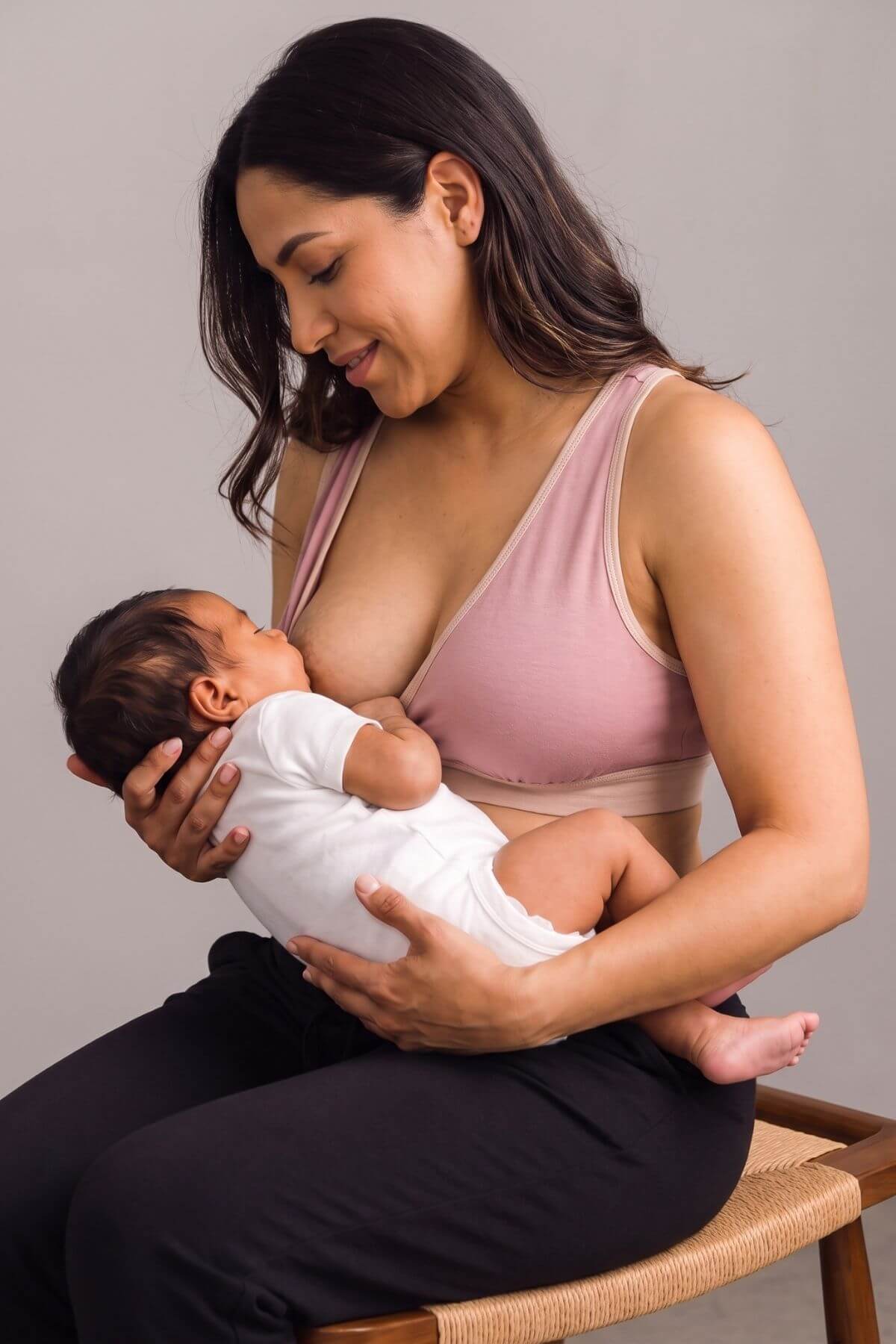 A woman in a pink nursing bra and black pants holds a baby wearing a white onesie while breastfeeding, seated on a wooden chair against a plain gray studio background.
