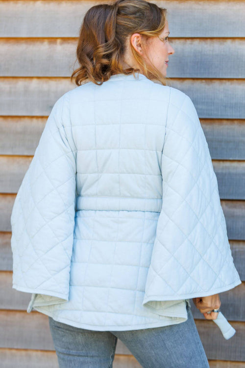 A woman viewed from the back wearing a light blue quilted jacket with wide bell sleeves, standing against a weathered wooden plank wall.#color_silver-cloud