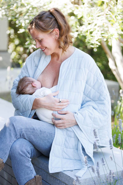 A woman sitting outdoors on a bench, wearing a light blue quilted baby blanket draped over her shoulders and across her lap as a nursing cover, with a baby nestled in her arms.