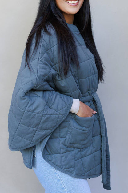 A woman wearing a slate blue-gray quilted jacket with a tie waist and front pockets, paired with light blue jeans, shown against a plain light gray background.