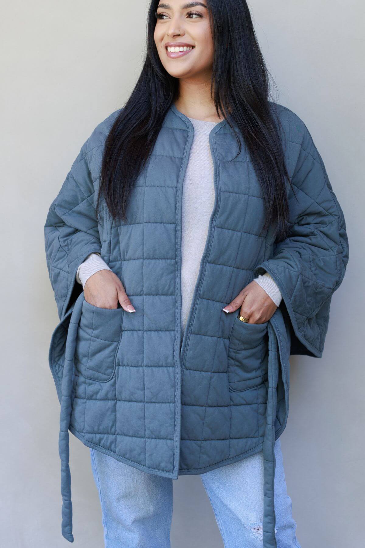 A woman wearing a slate blue-gray quilted jacket with a tie belt and front pockets, styled over a light gray top and light blue jeans, standing against a neutral gray wall.