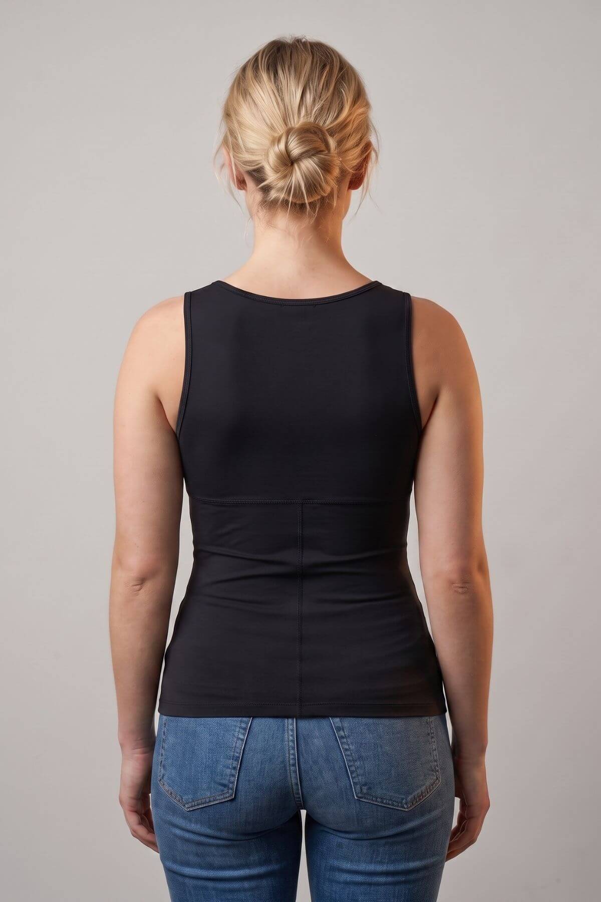 A woman viewed from the back wearing a black sleeveless tank top and blue jeans against a plain light gray studio background.