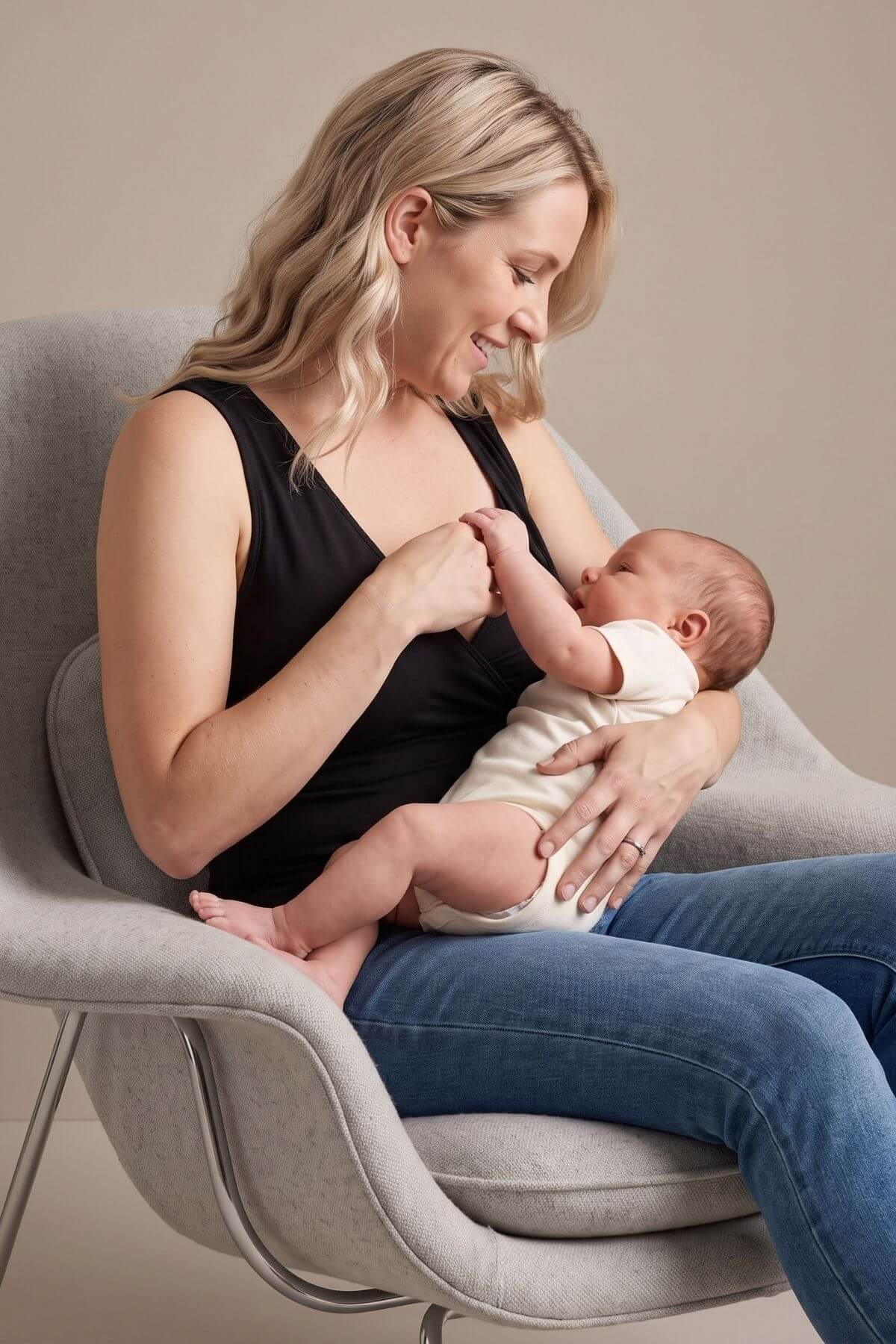 A woman in a black tank top and blue jeans sits in a light gray armchair, holding a baby dressed in a white outfit. The scene is photographed in a clean studio setting with a warm beige background.