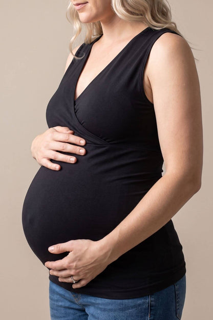 A pregnant woman wearing a black sleeveless wrap-style maternity top and blue jeans, shown from the side against a plain beige studio background.