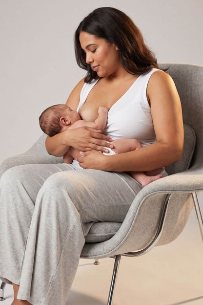 A woman in a white tank top and light gray sweatpants sits in a gray armchair, breastfeeding a baby. The background is a clean, neutral studio setting.