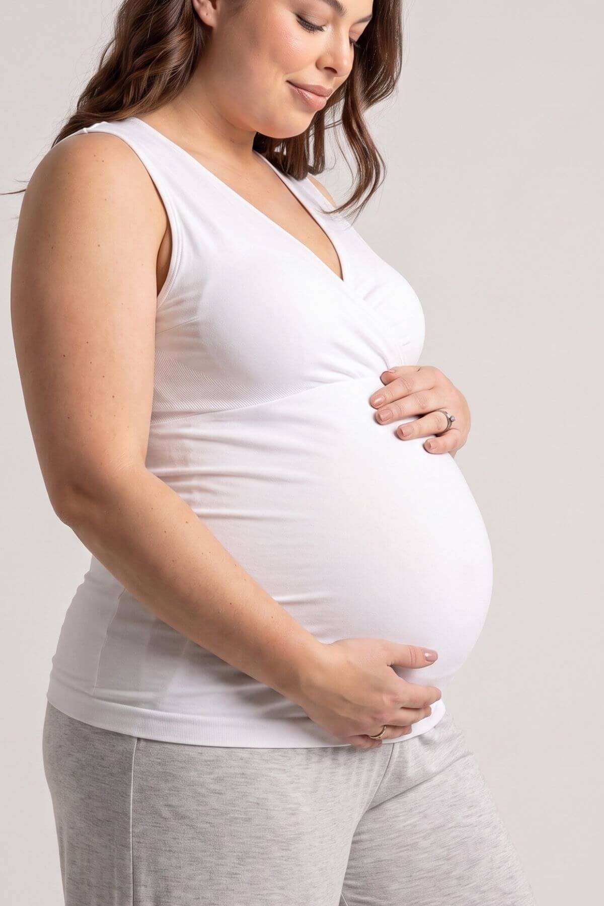 A pregnant woman wearing a white sleeveless V-neck maternity tank top and light gray sweatpants, holding her baby bump against a plain light gray studio background.