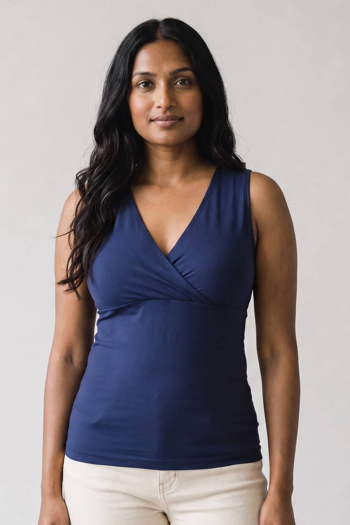 A woman modeling a navy blue sleeveless wrap-style top with a V-neckline, paired with light beige pants, against a plain light gray background.