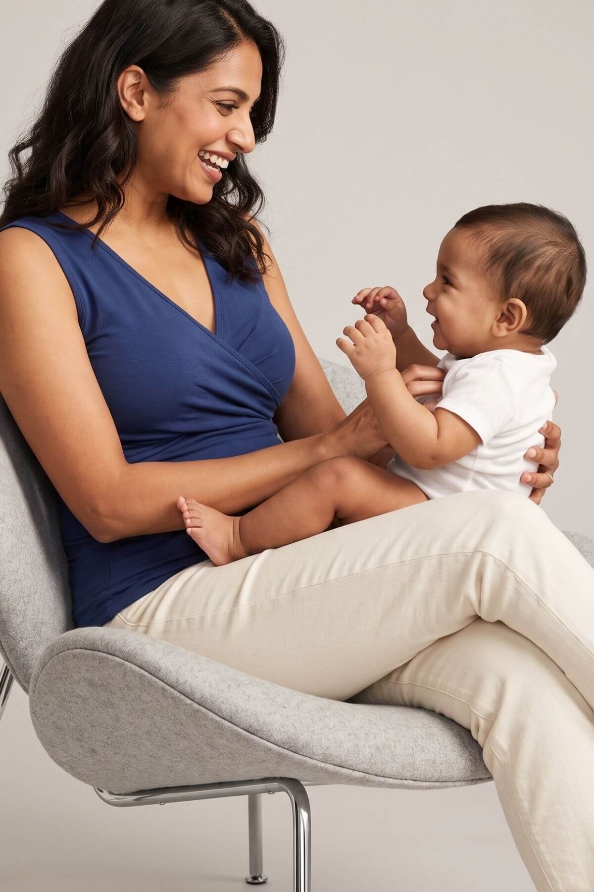 A woman in a navy blue sleeveless top and cream pants sits on a light gray upholstered chair, holding a smiling baby who is wearing a white onesie. The photo is shot in a clean studio setting with a neutral light gray background.