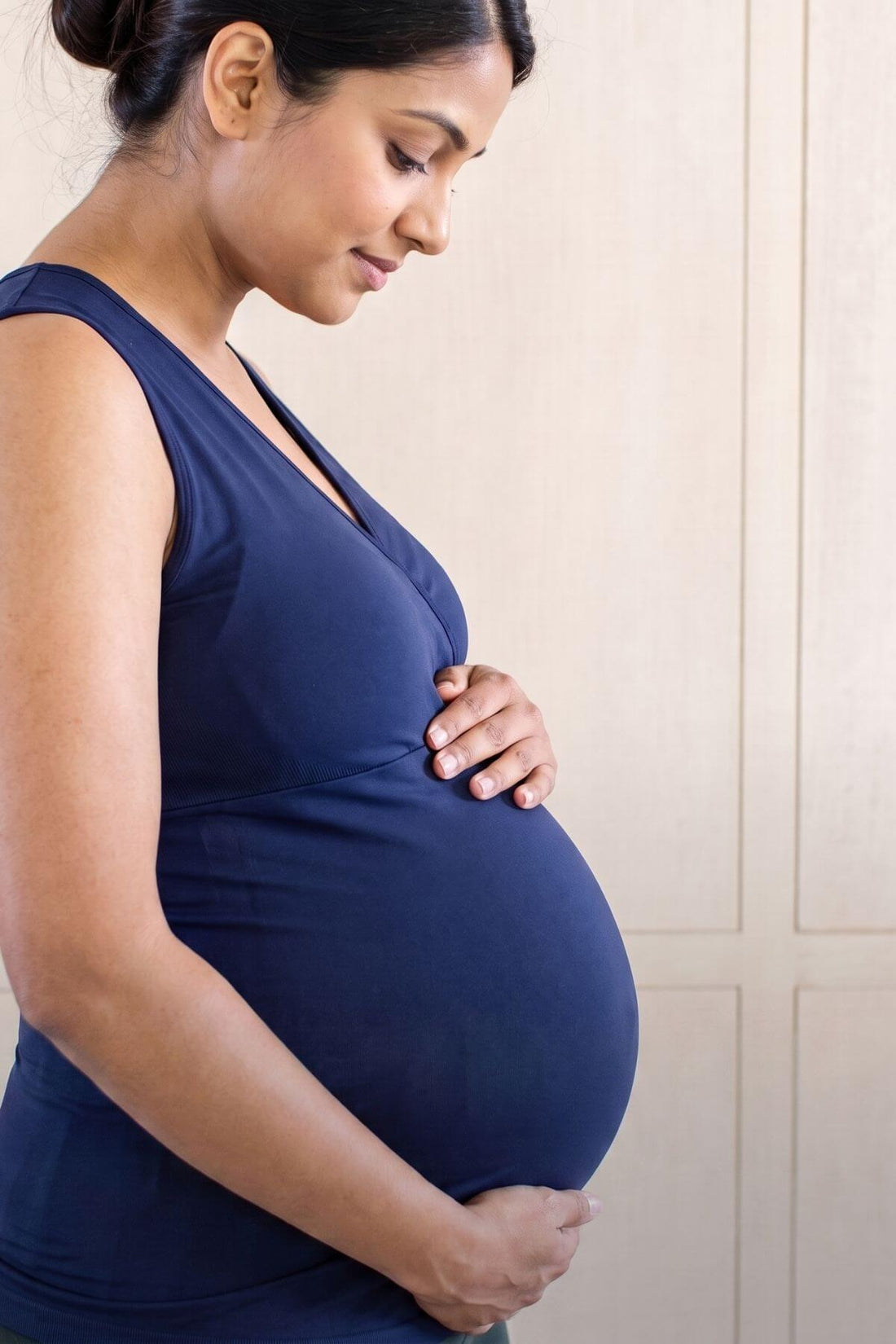 Pregnant woman in a navy blue sleeveless maternity dress, cradling her baby bump against a light beige paneled wall.