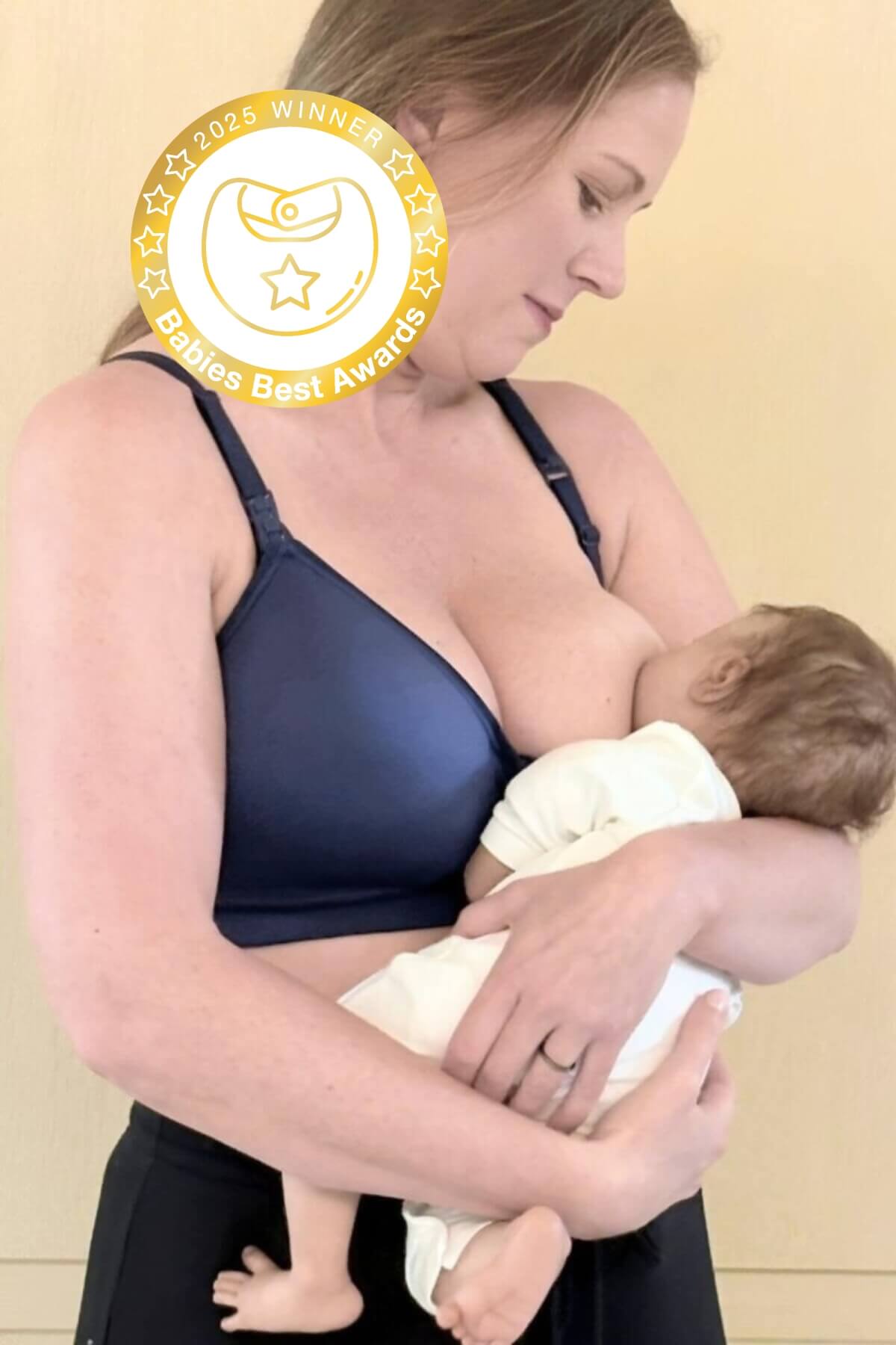 Woman wearing a hands free pumping and nursing bra whle breastfeeding a baby with an award badge in the corner