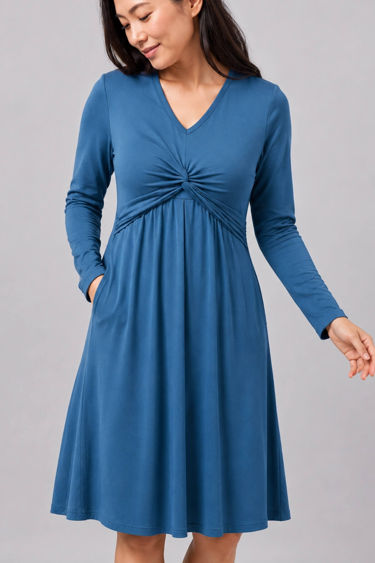 A woman wearing a long-sleeve blue V-neck dress with a gathered twist detail at the waist and side pockets, photographed against a plain light gray studio background