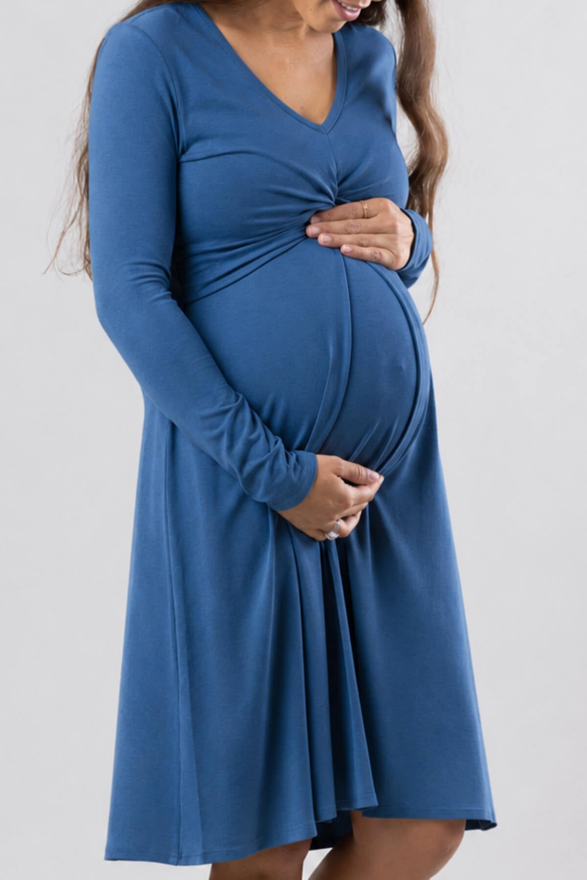 A pregnant woman wearing a long-sleeve blue maternity dress with a gathered twist detail at the bust, shown against a plain light gray background