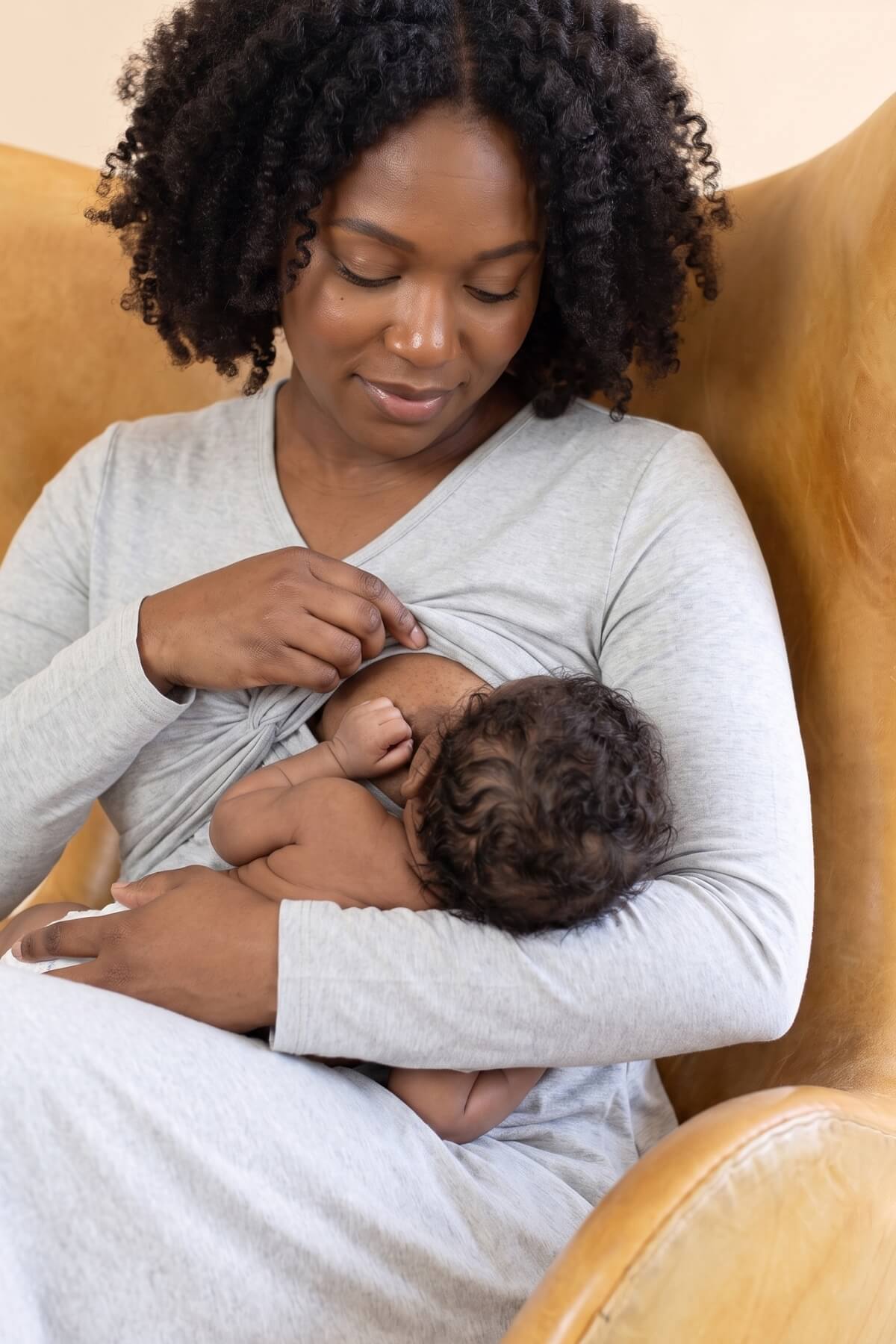 A woman with curly black hair wearing a light gray long-sleeve nursing top and matching bottoms sits in a tan leather armchair, breastfeeding a baby resting across her chest