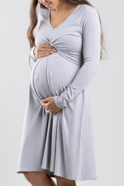 A pregnant woman wearing a light gray long-sleeve maternity dress, holding her baby bump against a plain light gray background