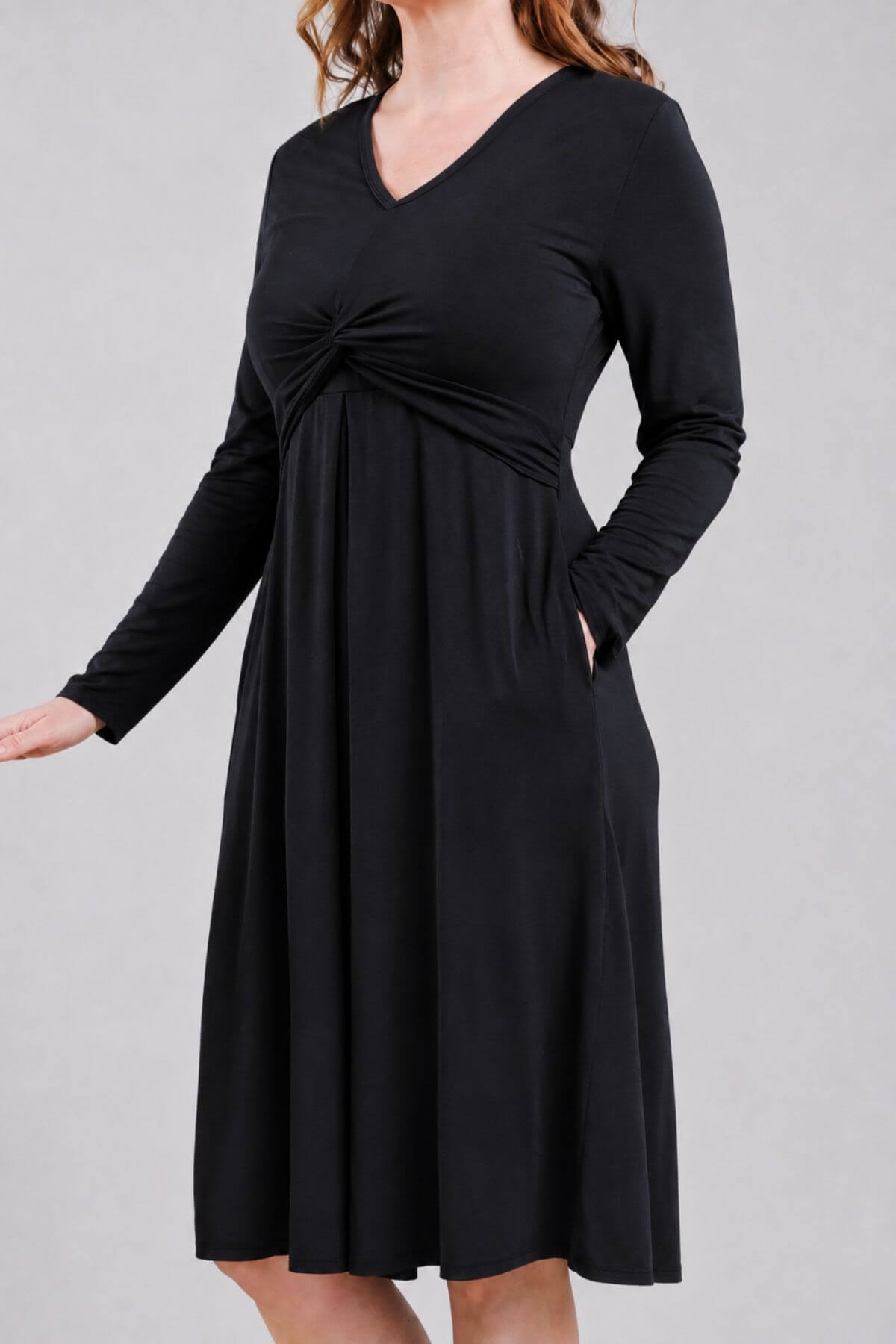 A woman wearing a black long-sleeve V-neck dress with a gathered twist detail at the waist and side pockets, shown against a plain light gray studio background