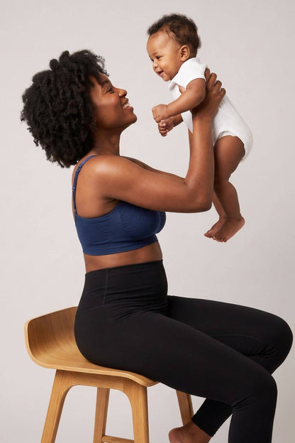 A woman sitting on a wooden stool holds a baby up against a light gray studio background. She is wearing the Stages Nursing Bra, Maternity Bra, Wearable Pump Bra and Everyday Bra all-in-one in indigo and black leggings.