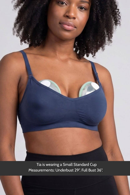 A woman wearing the Stages Nursing Bra, Maternity Bra, Wearable Pump Bra and Everyday Bra all-in-one in indigo with Elvie Stride wearable pumps inserted, shown from the chest up against a light gray background. A text banner at the bottom reads: &