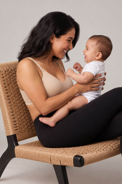 A woman sitting on a woven chair holds a smiling baby. She wears the Stages Maternity, Nursing, Wearable Pump &amp; Everyday Bra in Sunkissed Rose and black leggings, while the baby is in a white onesie.