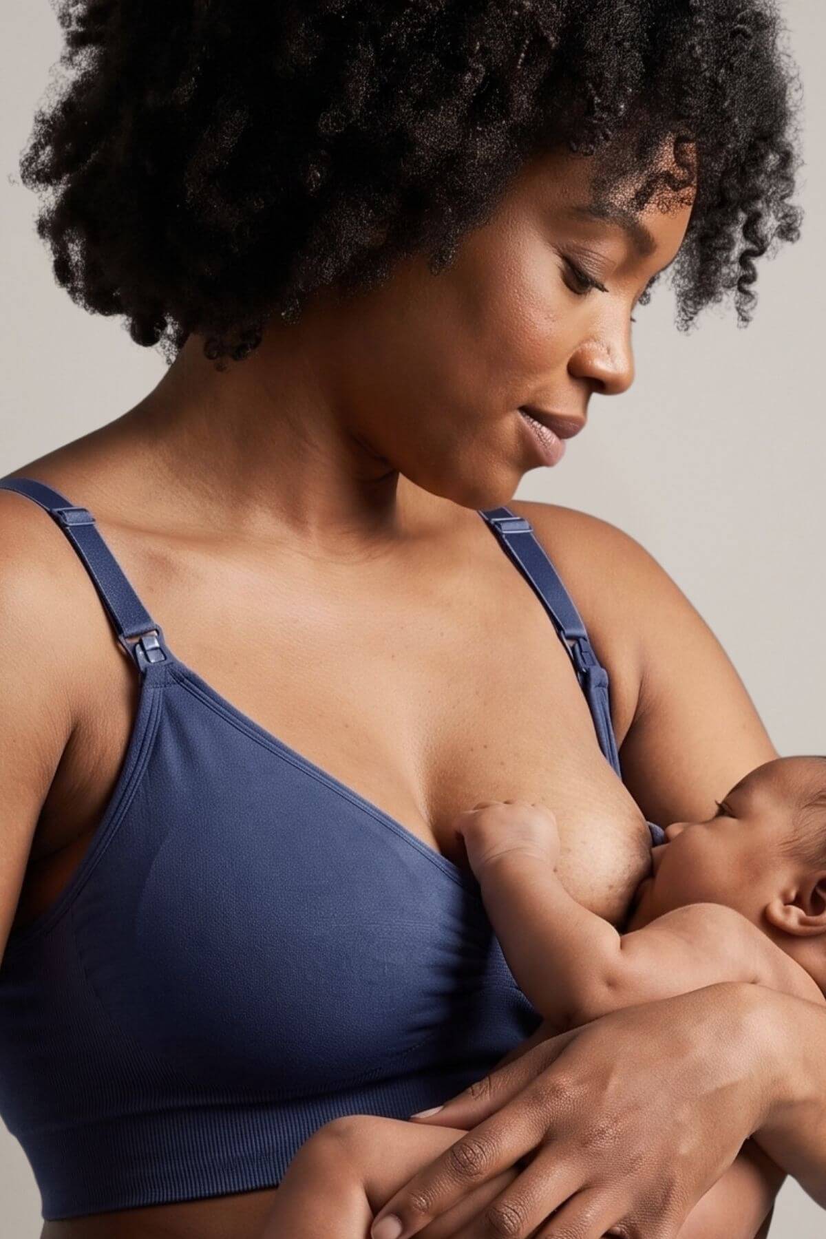 A close-up lifestyle shot of a woman with curly black hair breastfeeding a baby. She is wearing the Stages Nursing Bra, Maternity Bra, Wearable Pump Bra and Everyday Bra all-in-one in indigo against a neutral light gray background.