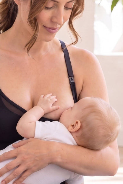 A woman wearing the Stages sweetheart mesh racerback style nursing bra  holds and breastfeeds a baby dressed in a white onesie in a bright, softly lit indoor setting.