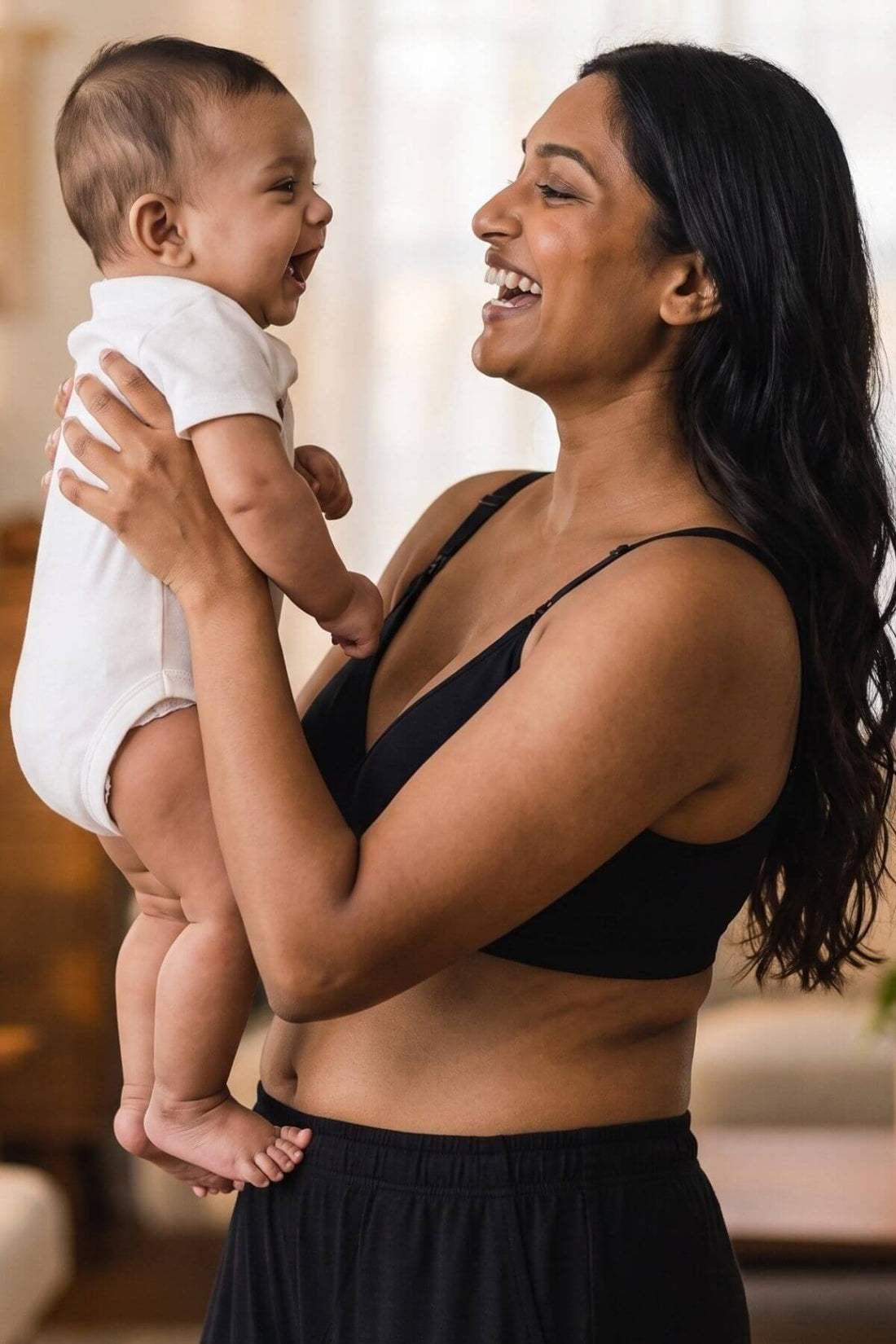A woman in a black simple wishes silhouette nursing &amp; hands free pumping bra and black pants holds a smiling baby dressed in a white onesie, set against a softly blurred indoor background.