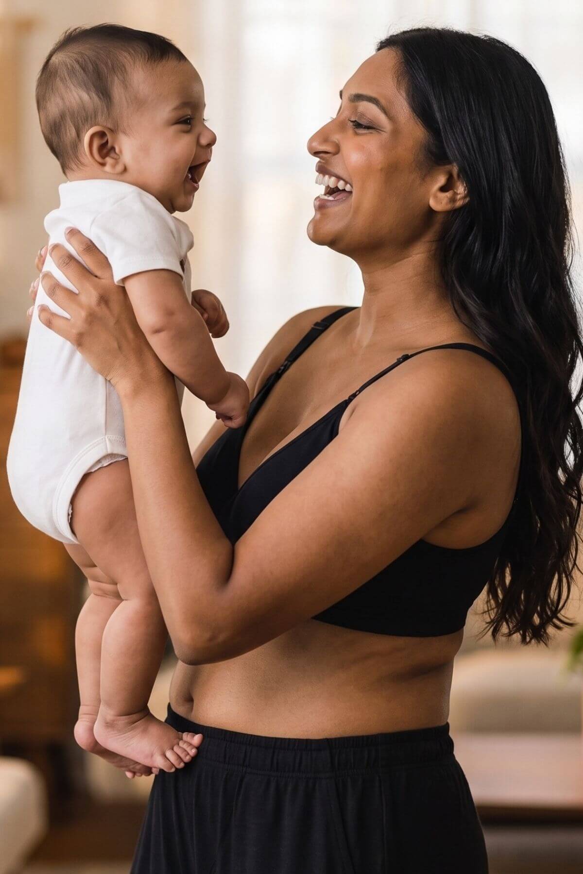 A woman in a black simple wishes silhouette nursing &amp; hands free pumping bra and black pants holds a smiling baby dressed in a white onesie, set against a softly blurred indoor background.