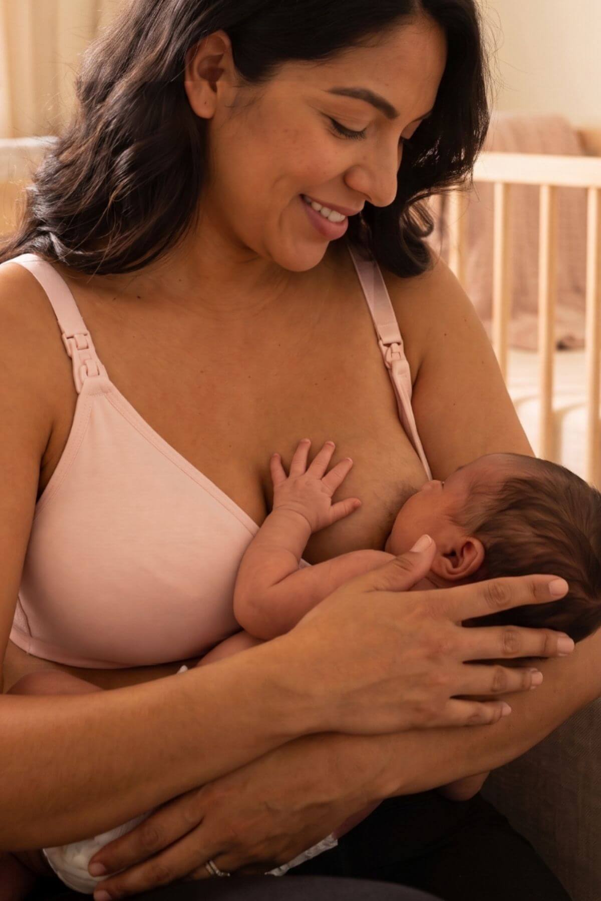 A woman wearing  a simple wishes supermom silhouette nursing bra and hands free pumping bra in ballet slipper pink holds and breastfeeds a newborn baby in a warm, softly lit indoor setting.