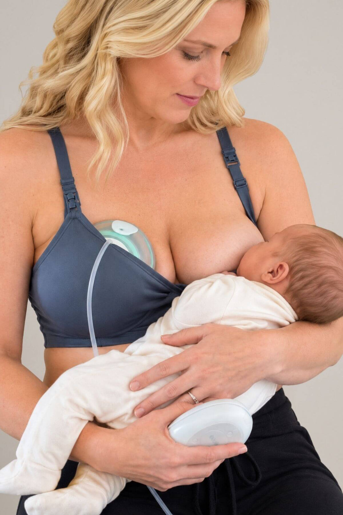 A woman breastfeeding a baby while wearing a dark blue wireless nursing bra and a white feeding pump connected to a clear tube. The baby is dressed in a white onesie.