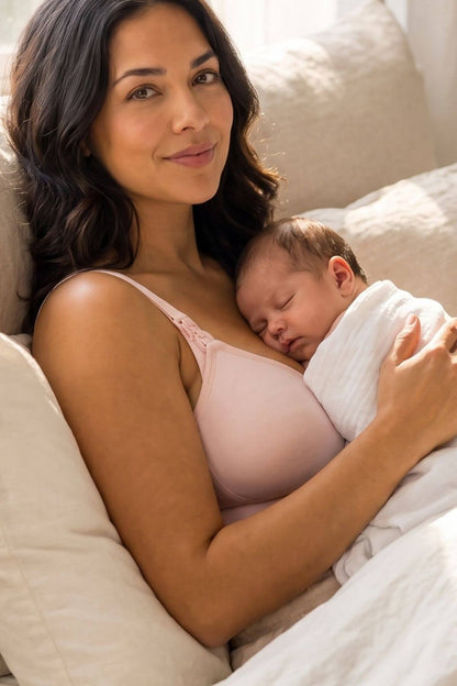 A woman in a simple wishes supermom silhouette nursing bra and hands free pumping bra in ballet slipper pink breastfeeding a sleeping newborn baby wrapped in a white blanket, resting on beige pillows.