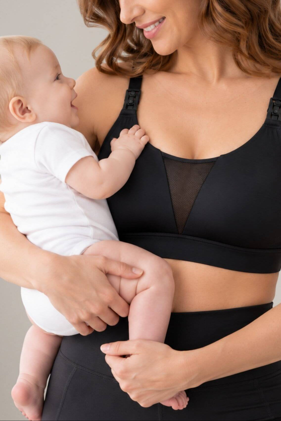 A woman wearing the Supermom V mesh tank style nursing and hands free pumping sports bra  and matching bottoms holds a baby dressed in a white onesie against a plain light gray background.