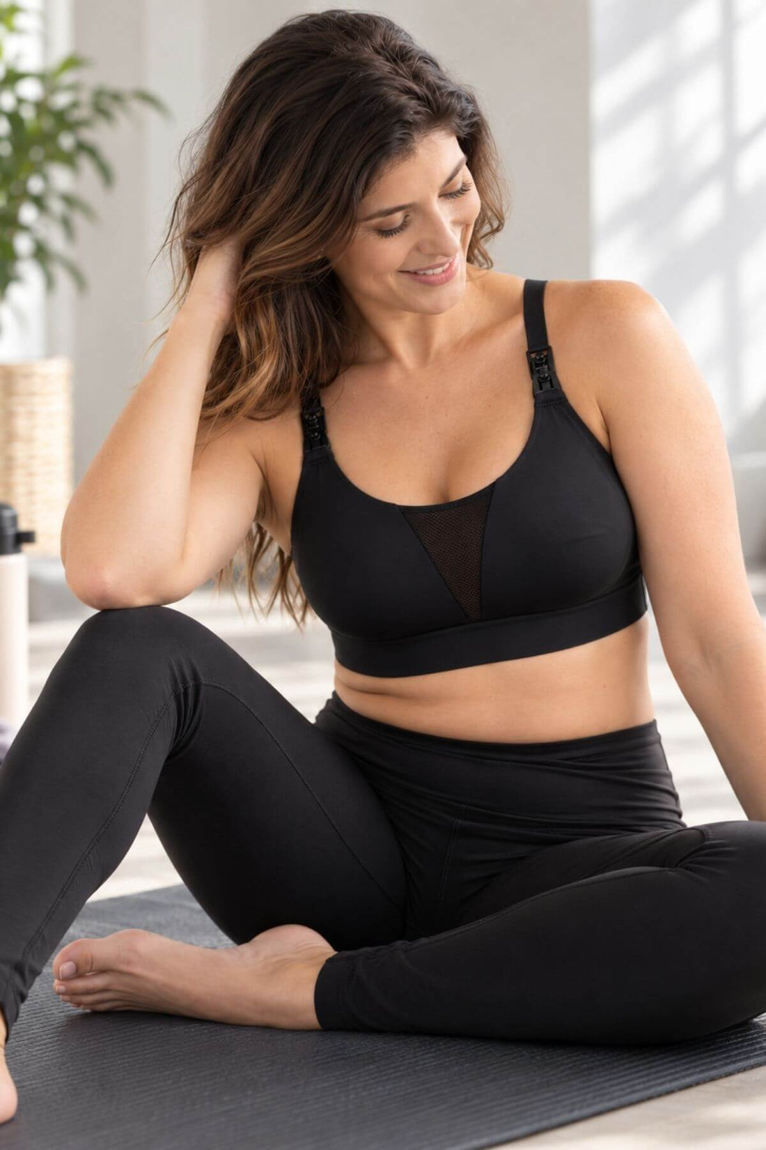 A woman sits cross-legged on a yoga mat wearing the Supermom V mesh tank style nursing and hands free pumping sports bra and matching black leggings in a bright indoor setting.