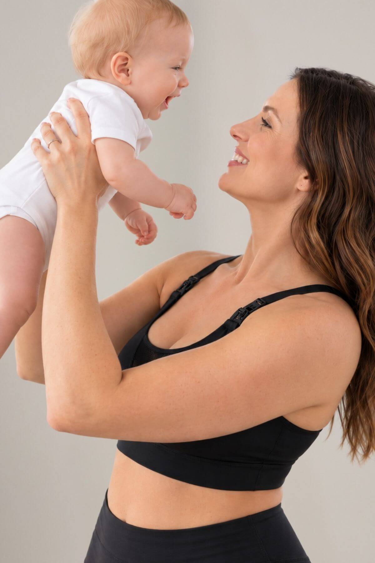 A woman in the Stages v-mesh inset tank style nursing and wearable pump support bra  and matching bottoms holds a smiling baby wearing a white onesie against a plain light gray background.