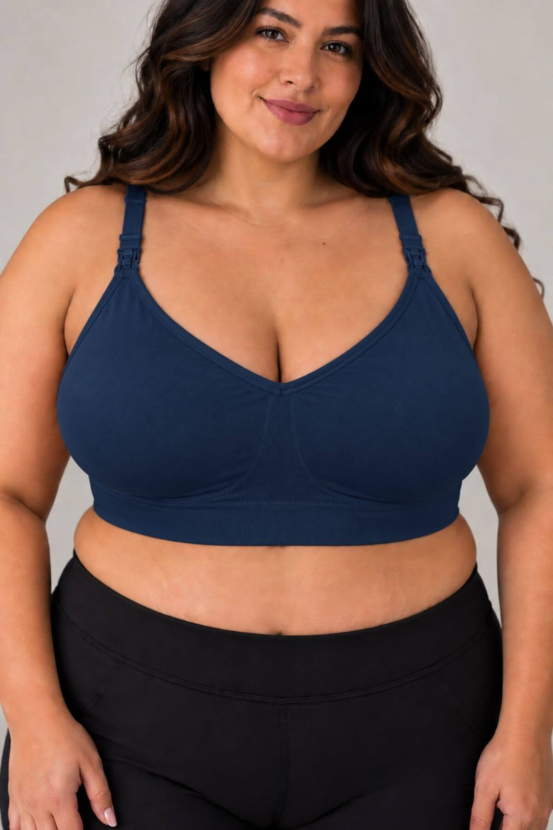 A plus size woman wearing the supermom silhouette hands free pumping & nursing bra in indigo and black leggings, shown from the chest down against a plain light gray studio background.