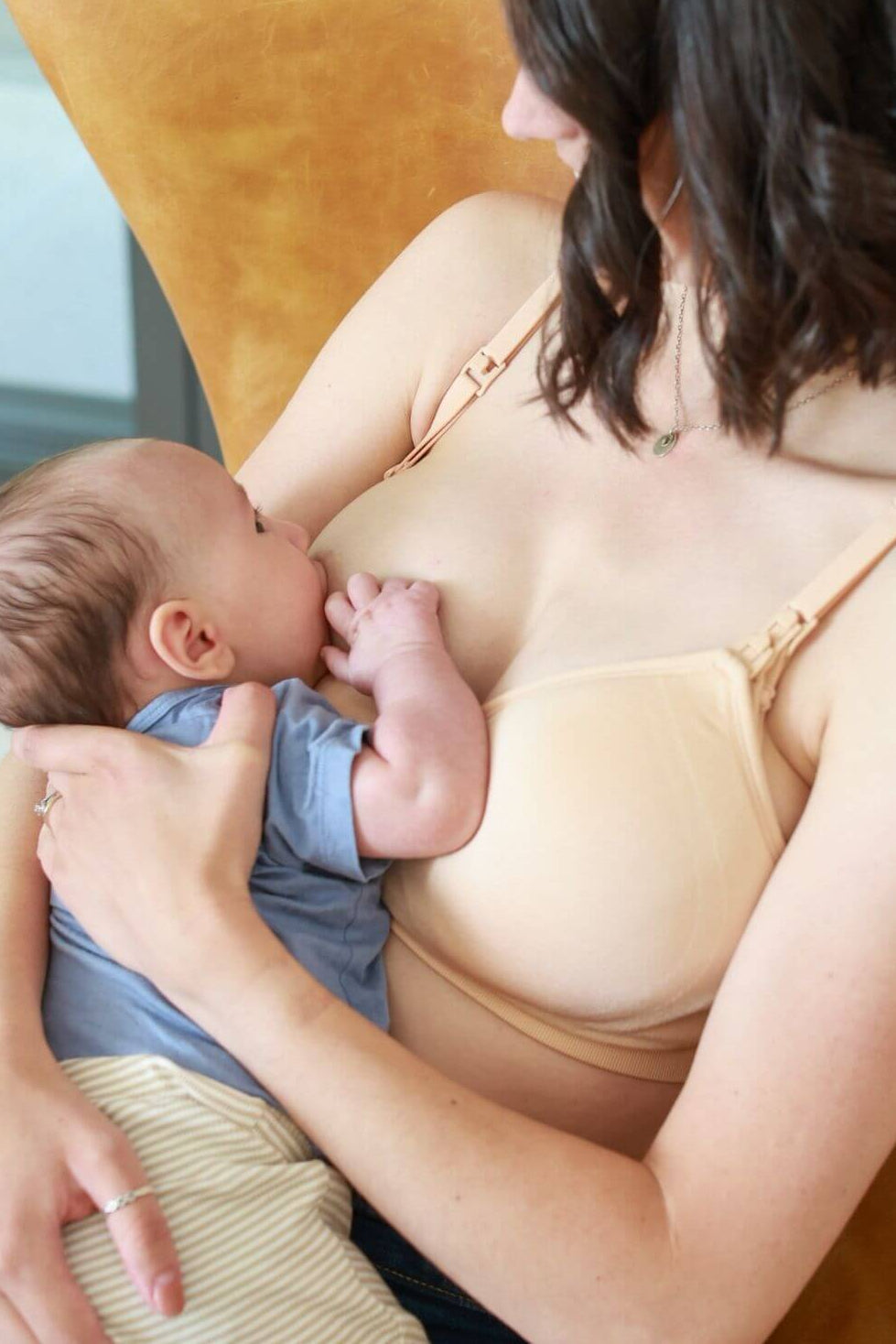 A woman in the supermom silhouette nursing and hands free pumping bra in beige holds a baby while breastfeeding, seated in a mustard chair. A text banner at the bottom reads: &