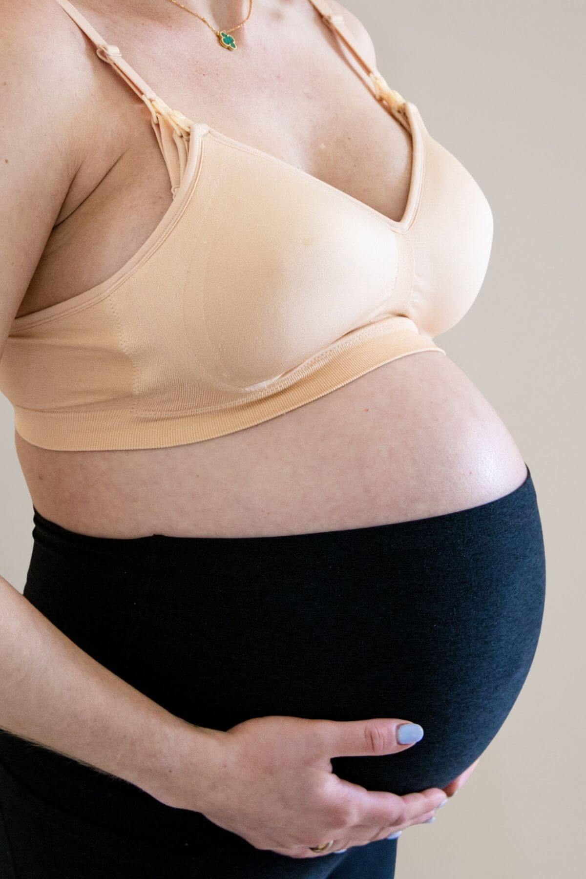 Pregnant woman wearing the simple wishes supermom silhouette nursing and hands free pumping bra in beige and black maternity belly band, holding her baby bump against a neutral beige background.