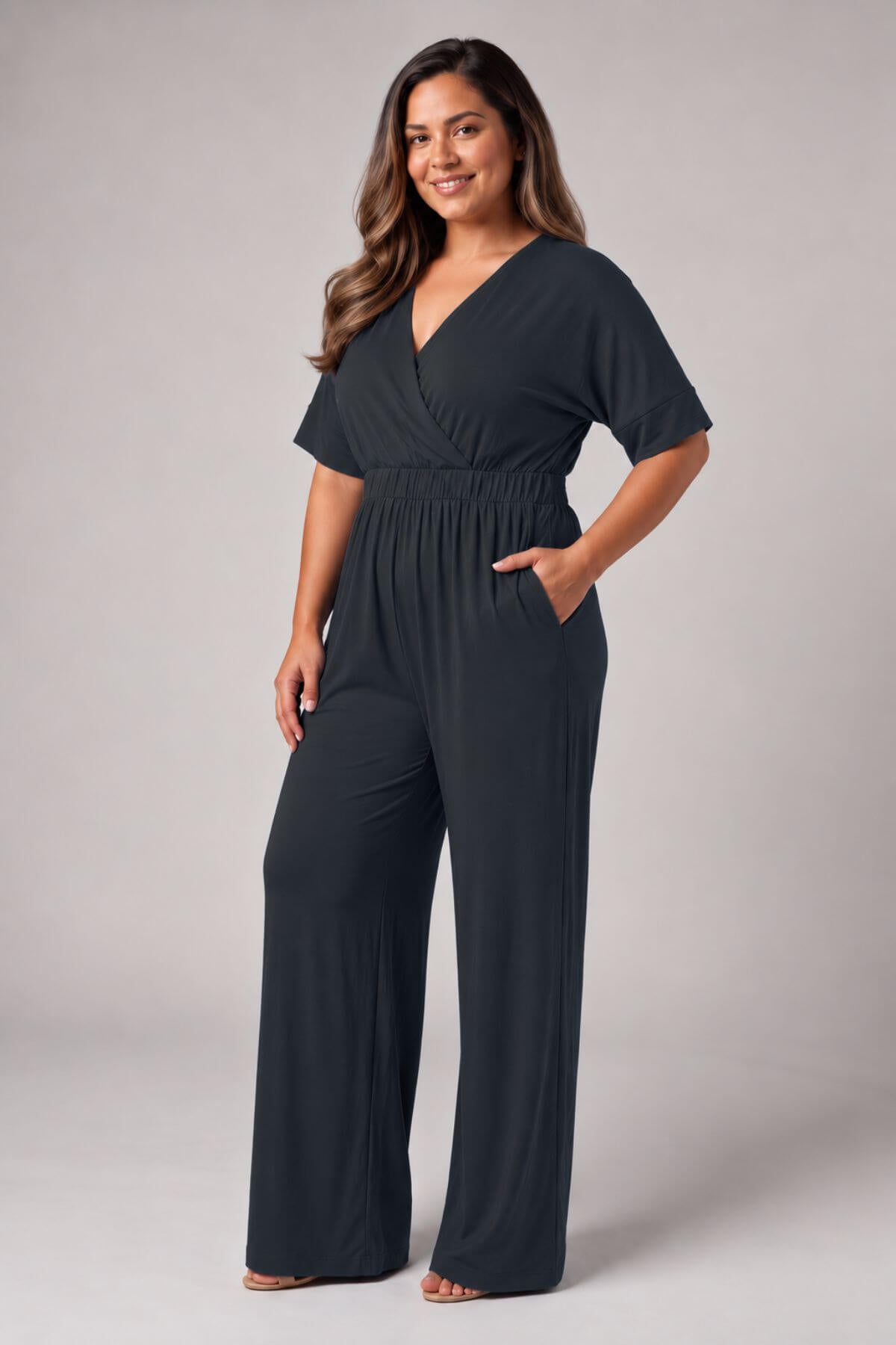 A woman modeling a dark charcoal gray jumpsuit with a V-neckline, short sleeves, a smocked elastic waist, and wide legs, set against a neutral studio background.