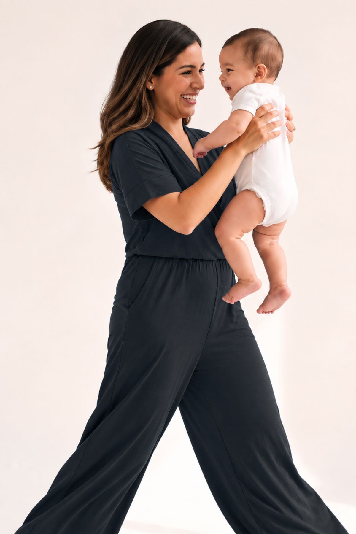 A woman in a dark charcoal gray short-sleeve jumpsuit holds a baby wearing a white bodysuit against a plain light background.