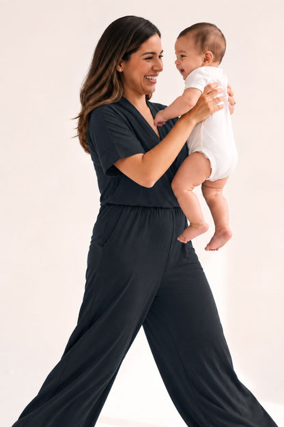A woman in a dark charcoal gray short-sleeve jumpsuit holds a baby wearing a white bodysuit against a plain light background.