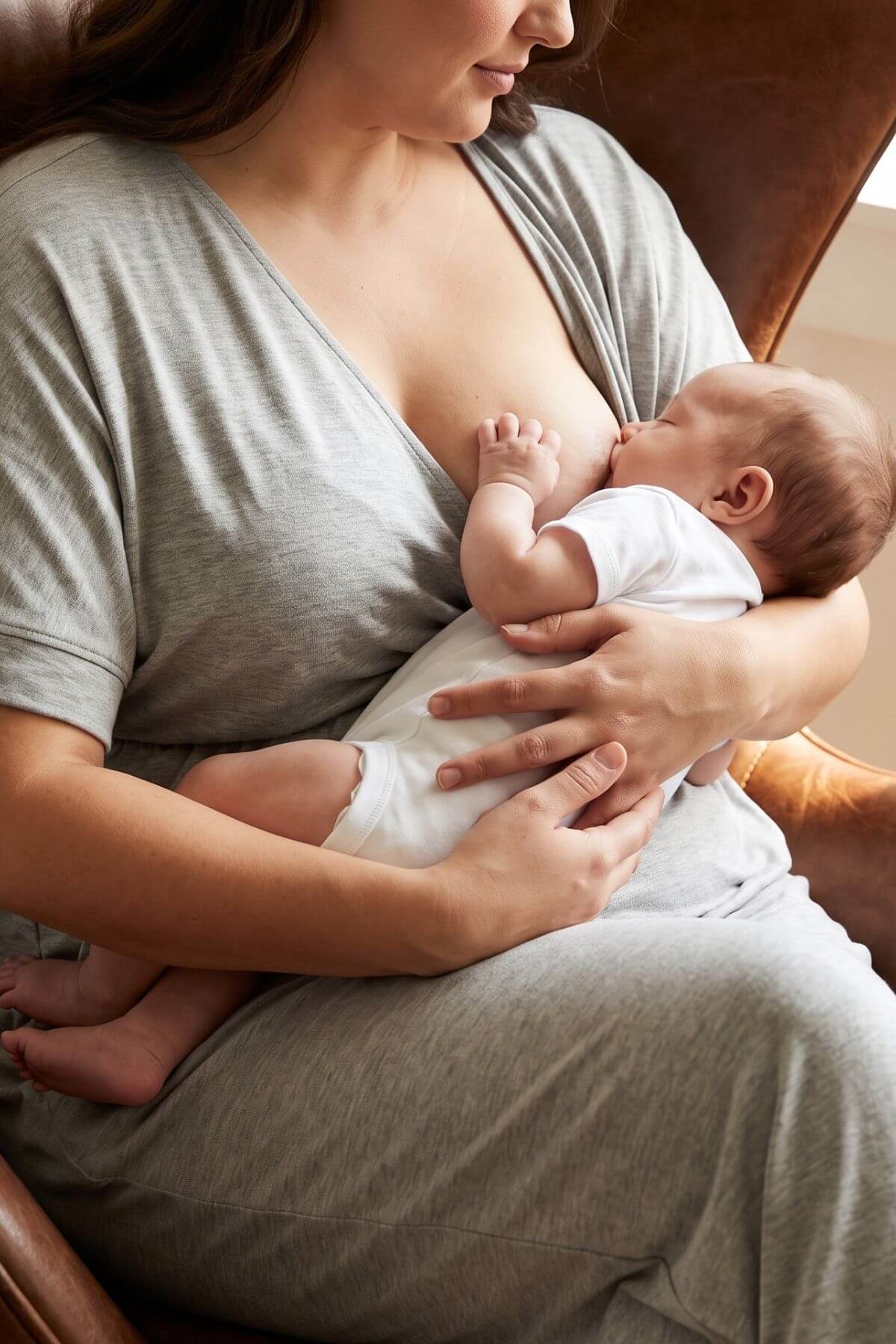 A woman in a gray lounge set holds a baby wearing a white bodysuit while breastfeeding, seated in a brown leather armchair.