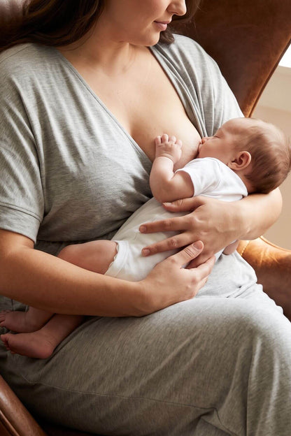 A woman in a gray lounge set holds a baby wearing a white bodysuit while breastfeeding, seated in a brown leather armchair.