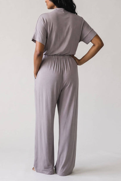 A woman shown from the back wearing a matching short-sleeve top and wide-leg pants set in a dusty mauve-gray color, set against a plain light gray studio background..