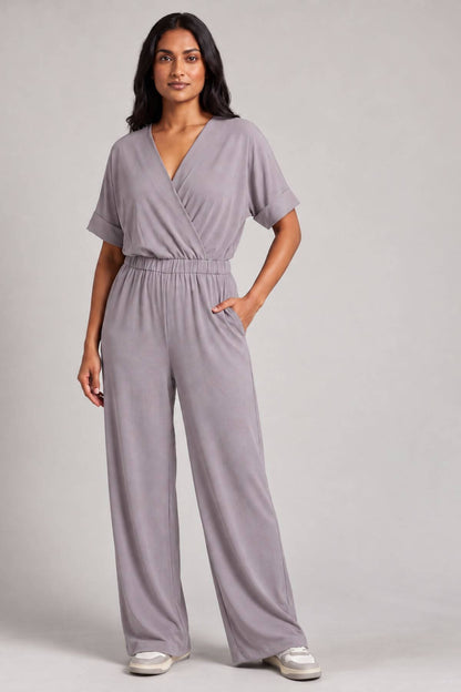 A woman modeling a light purple-gray short-sleeve wrap jumpsuit with wide legs, standing against a neutral light gray studio background..