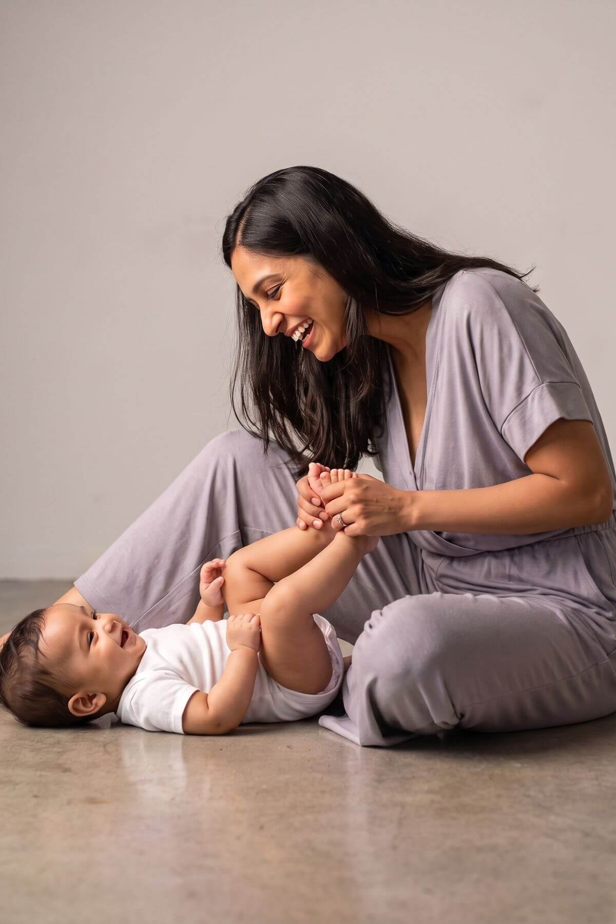 A woman in a matching light purple loungewear set sits on a concrete floor, smiling as she playfully lifts a baby&